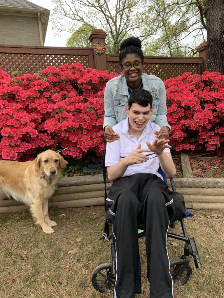 A caregiver rests her hands on the shoulders of a smiling young man in his wheelchair in a backyard garden, next to a golden retriever